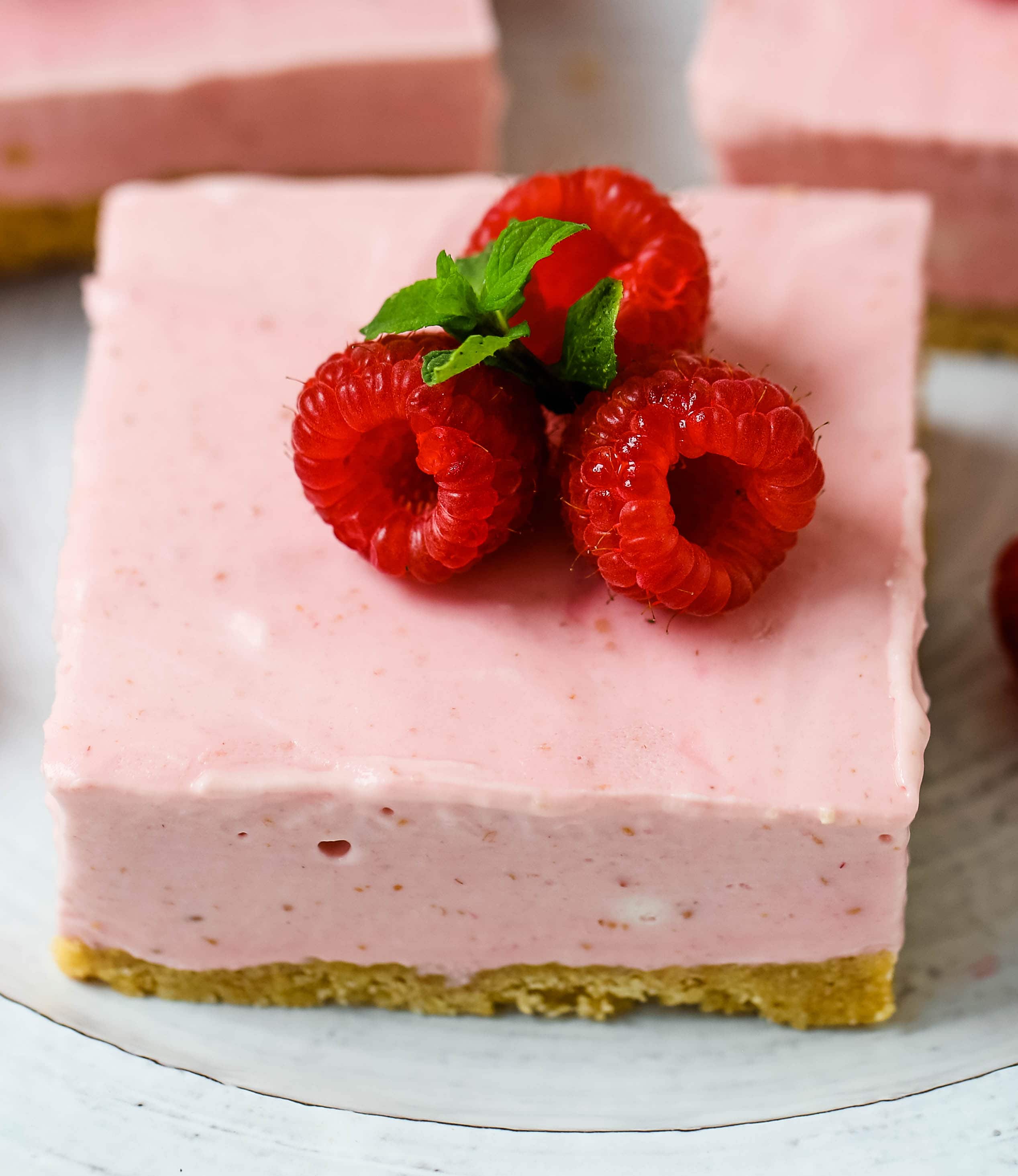 No-Bake Frozen Raspberry Cheesecake Squares. Creamy no-bake raspberry cheesecake filling on top of a shortbread or graham cracker crust topped with fresh raspberries. A creamy no-bake frozen dessert.  www.modernhoney.com #nobakedessert #raspberrycheesecake #cheesecakesquares #frozendessert #frozencheesecake