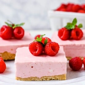 No-Bake Frozen Raspberry Cheesecake Squares. Creamy no-bake raspberry cheesecake filling on top of a shortbread or graham cracker crust topped with fresh raspberries. A creamy no-bake frozen dessert. www.modernhoney.com #nobakedessert #raspberrycheesecake #cheesecakesquares #frozendessert #frozencheesecake