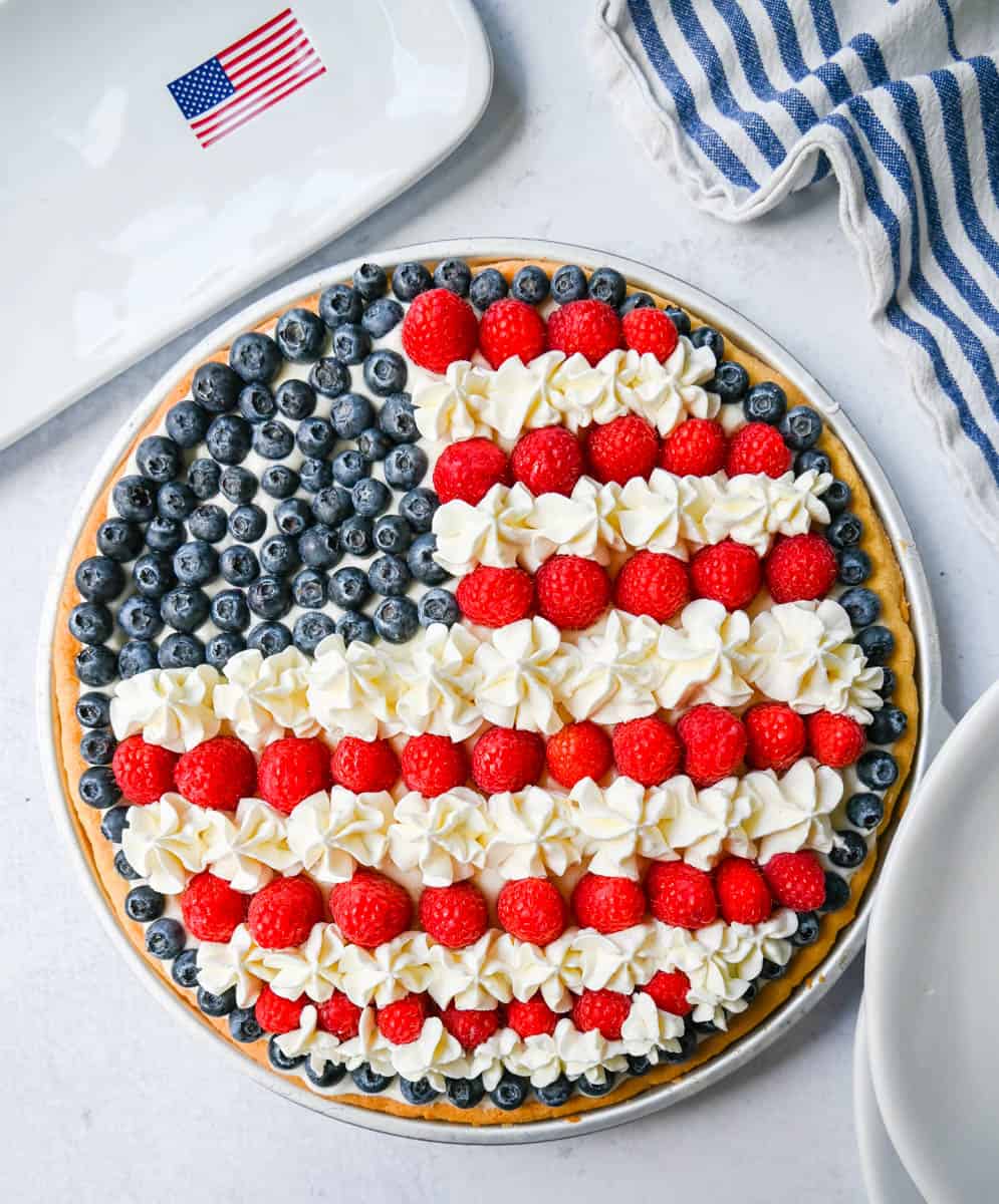 This flag sugar cookie fruit pizza is a festive, delicious, and beautiful patriotic 4th of July dessert featuring a homemade soft sugar cookie crust with a sweet vanilla cream cheese frosting topped with fresh raspberries, blueberries, and homemade whipped cream.