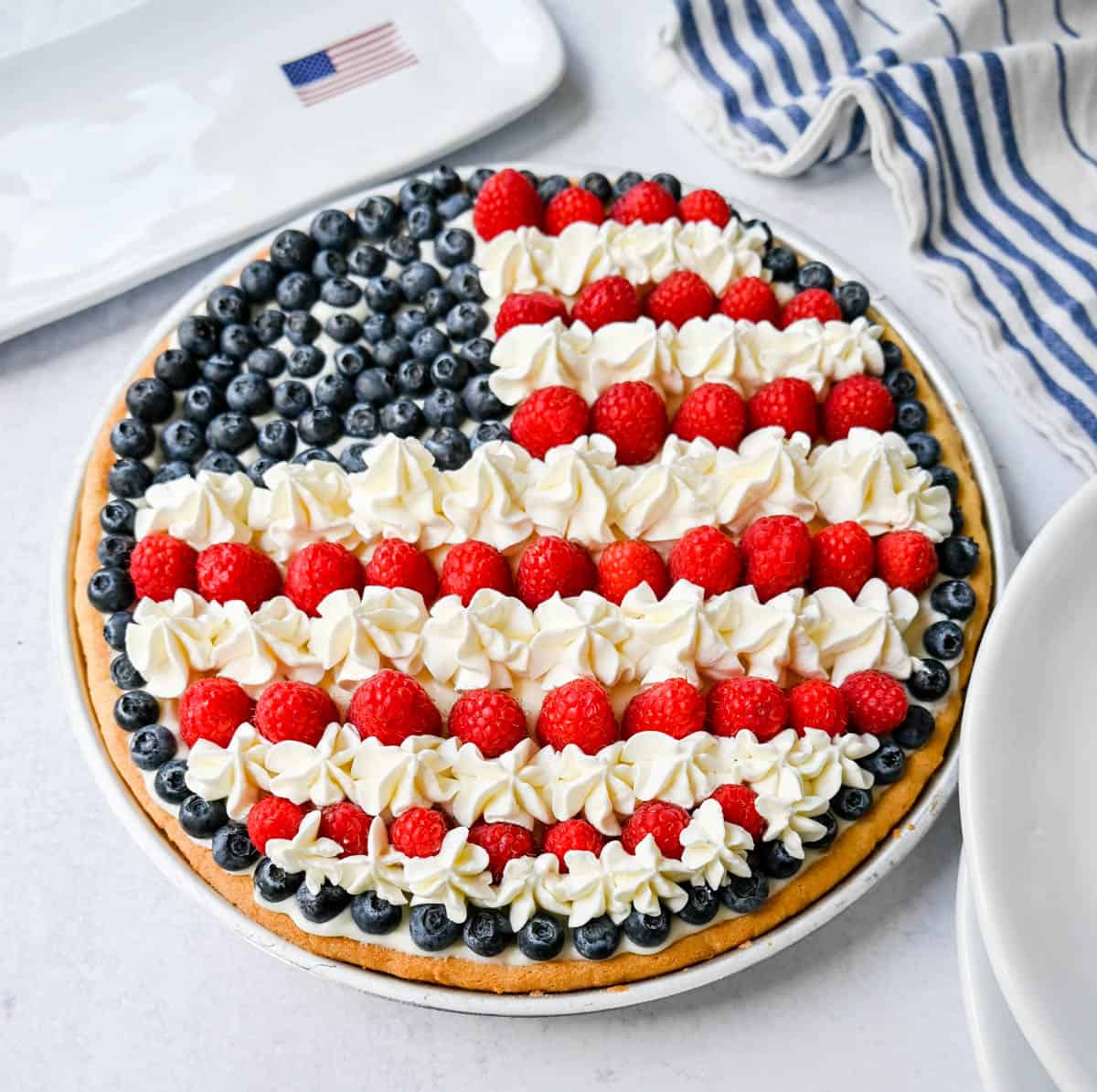 This flag sugar cookie fruit pizza is a festive, delicious, and beautiful patriotic 4th of July dessert featuring a homemade soft sugar cookie crust with a sweet vanilla cream cheese frosting topped with fresh raspberries, blueberries, and homemade whipped cream. 