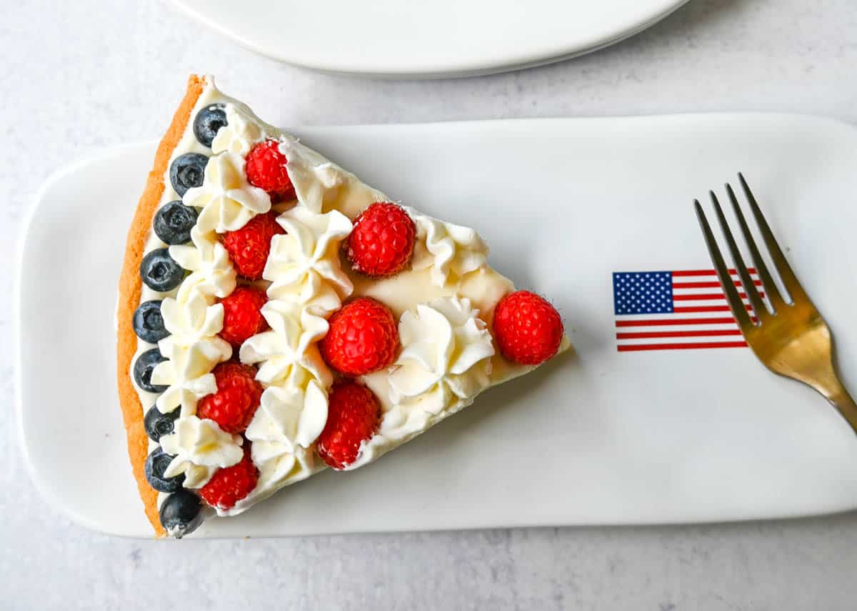This flag sugar cookie fruit pizza is a festive, delicious, and beautiful patriotic 4th of July dessert featuring a homemade soft sugar cookie crust with a sweet vanilla cream cheese frosting topped with fresh raspberries, blueberries, and homemade whipped cream. 