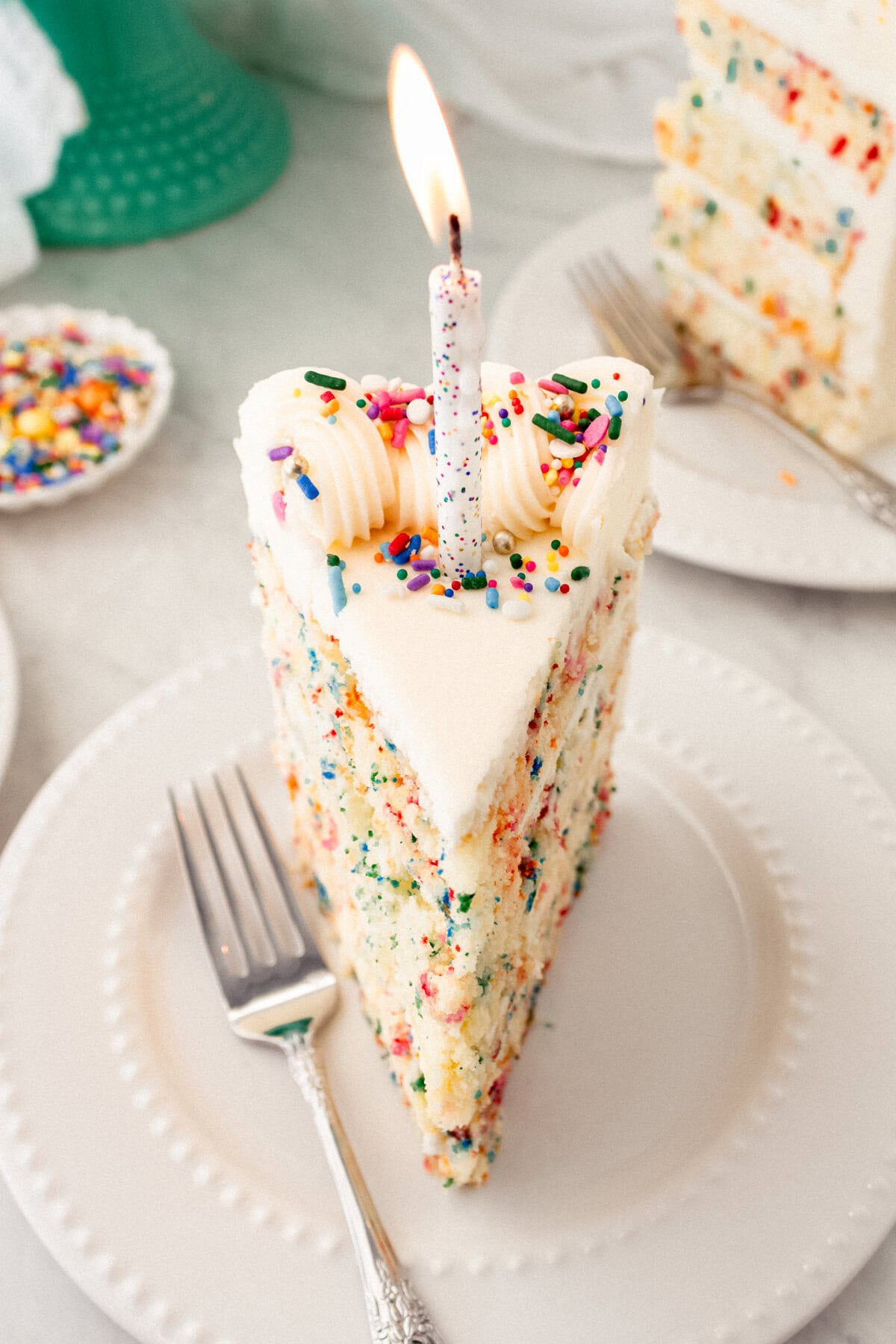 Piece of birthday cake with sprinkles with a candle. A fluffy homemade Funfetti birthday cake with rainbow sprinkles topped with a light cream cheese buttercream frosting and an array of sprinkles. This Confetti Cake recipe is the perfect birthday cake!