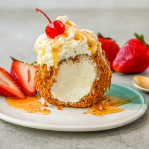 Crunchy cinnamon-sugar corn flakes coat creamy scoops of ice cream, topped with honey, whipped cream, and strawberries. This No-Fry Fried Ice Cream has zero deep-frying involved. This easy fried ice cream recipe uses corn flakes coated in buttery cinnamon sugar to mimic the crunch of a fried shell. No hot oil, no mess — just roll, freeze, and serve.
