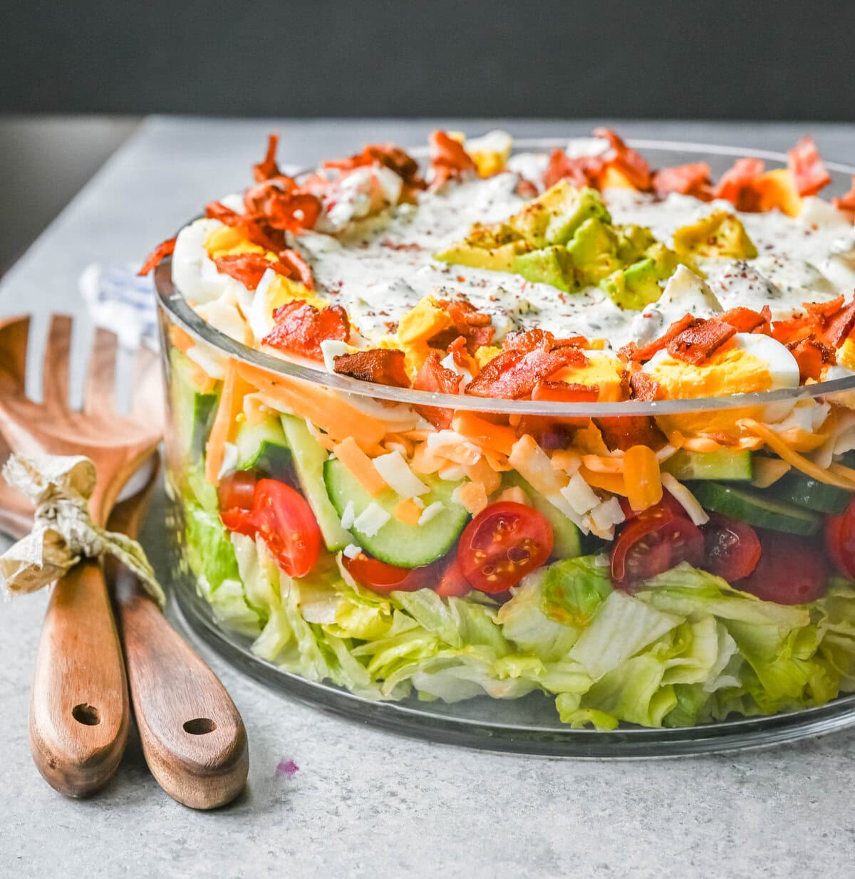This classic Seven Layer salad is layered with iceberg lettuce, smoky bacon, hard-boiled eggs, sharp cheddar cheese, fresh veggies, and your choice of peas or garbanzo beans, plus creamy avocado. It's the ultimate make-ahead side dish, perfect for BBQs or family dinners. This easy, crowd-pleasing 7 Layer Salad is topped with a generous drizzle of homemade ranch dressing. This easy, crowd-pleasing layered salad is a beautiful, delicious side dish!