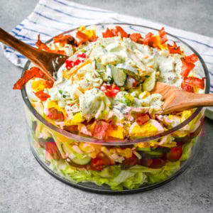 This classic Seven Layer salad is layered with iceberg lettuce, smoky bacon, hard-boiled eggs, sharp cheddar cheese, fresh veggies, and your choice of peas or garbanzo beans, plus creamy avocado. It's the ultimate make-ahead side dish, perfect for BBQs or family dinners. This easy, crowd-pleasing 7 Layer Salad is topped with a generous drizzle of homemade ranch dressing. This easy, crowd-pleasing layered salad is a beautiful, delicious side dish!