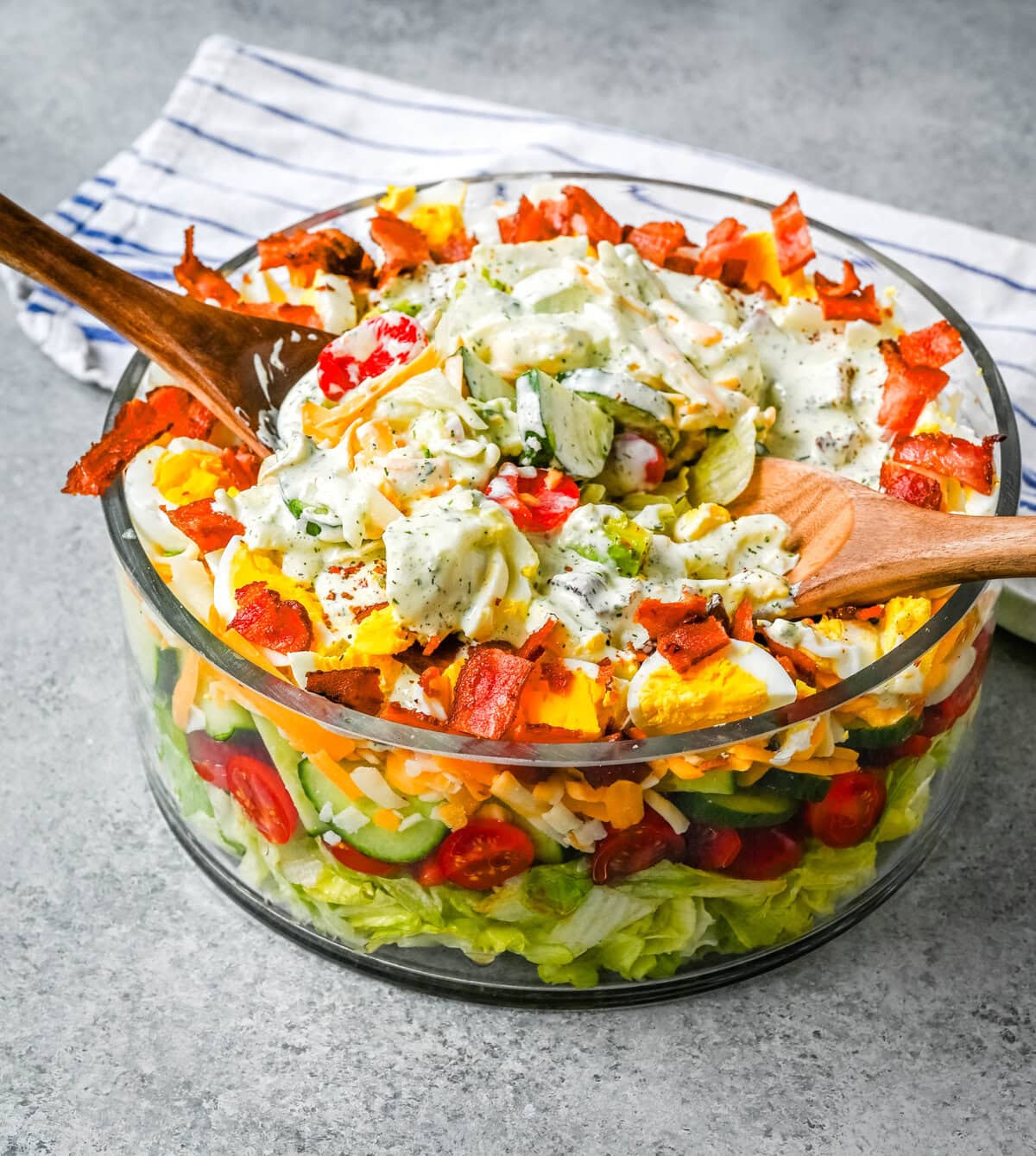 This classic Seven Layer salad is layered with iceberg lettuce, smoky bacon, hard-boiled eggs, sharp cheddar cheese, fresh veggies, and your choice of peas or garbanzo beans, plus creamy avocado. It's the ultimate make-ahead side dish, perfect for BBQs or family dinners. This easy, crowd-pleasing 7 Layer Salad is topped with a generous drizzle of homemade ranch dressing. This easy, crowd-pleasing layered salad is a beautiful, delicious side dish!