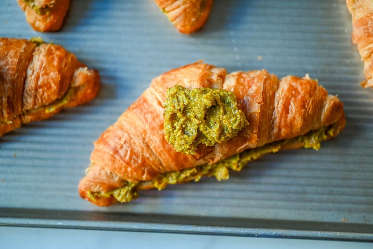 Fill the croissant with homemade pistachio fillng and then top with a dollop of pistachio filling on the top of the croissant before baking.